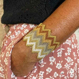 May include: A gold, gray, and brown chevron patterned bracelet. The bracelet is made of woven thread and is worn on a person's wrist.