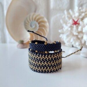 May include: Three black and gold friendship bracelets with gold beads. The bracelets are stacked on top of each other.