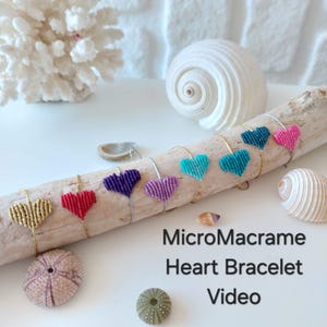 以下が含まれることがあります： 流木に飾られた、カラフルなマイクロマクラメのハートブレスレットのコレクション。ブレスレットは、金、赤、紫、薄紫、ターコイズ、濃いターコイズ、ピンクのハートが特徴です。画像には貝殻と「MicroMacrame Heart Bracelet Video」の文字も含まれています。