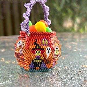 Puede incluir: Una peque&ntilde;a cesta redonda hecha de cuentas con tem&aacute;tica de Halloween, que presenta un patr&oacute;n de bruja, fantasma y casa en naranja, negro y morado. La cesta tiene un asa de cuentas color lavanda con volantes y est&aacute; llena de objetos redondos.