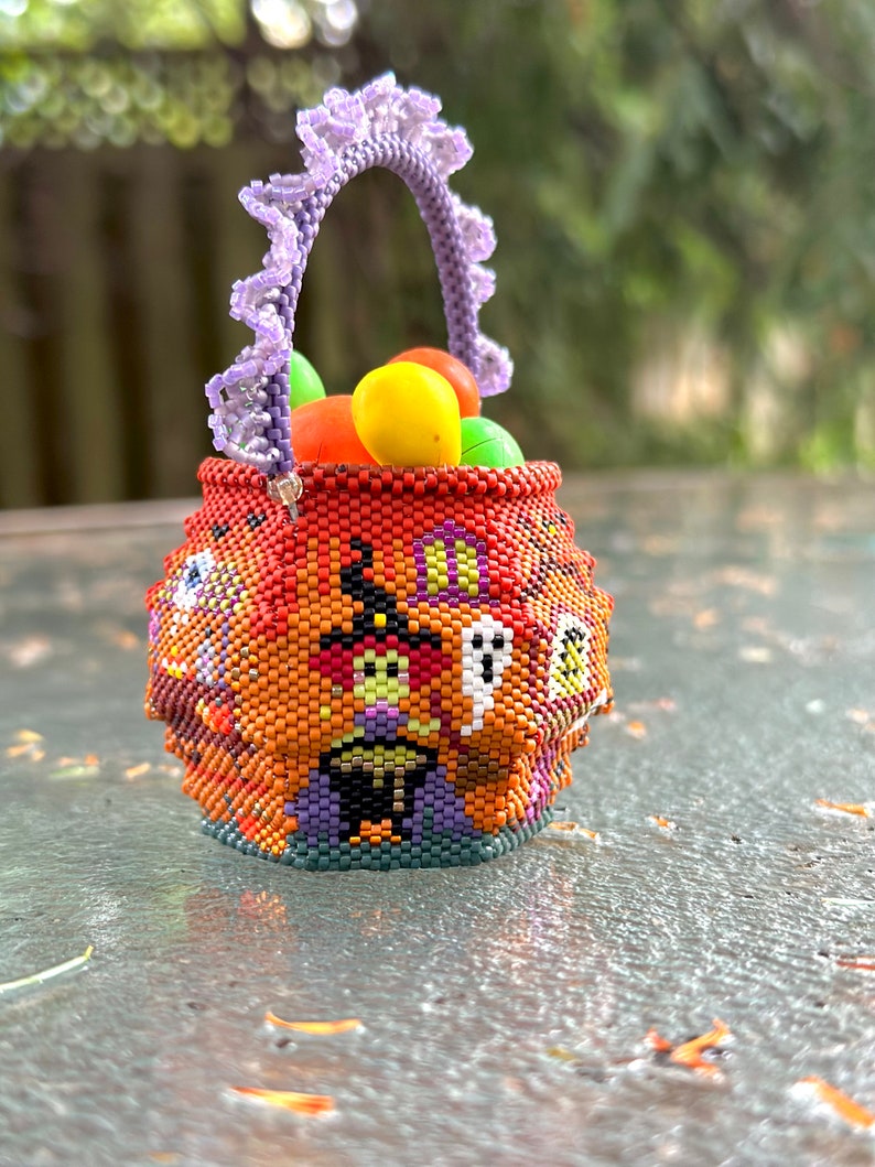 Puede incluir: Una peque&ntilde;a cesta redonda hecha de cuentas con tem&aacute;tica de Halloween, que presenta un patr&oacute;n de bruja, fantasma y casa en naranja, negro y morado. La cesta tiene un asa de cuentas color lavanda con volantes y est&aacute; llena de objetos redondos.
