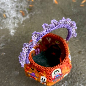 Puede incluir: Una peque&ntilde;a cesta redonda de cuentas con un exterior naranja que presenta un patr&oacute;n repetitivo de fantasmas blancos y motivos coloridos. La cesta tiene un asa curva adornada con un borde de cuentas de color violeta claro con volantes.