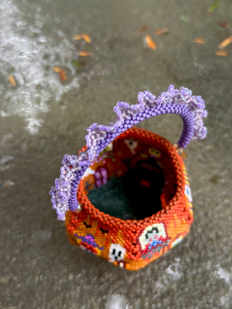 Puede incluir: Una peque&ntilde;a cesta redonda de cuentas con un exterior naranja que presenta un patr&oacute;n repetitivo de fantasmas blancos y motivos coloridos. La cesta tiene un asa curva adornada con un borde de cuentas de color violeta claro con volantes.