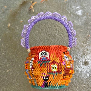 Puede incluir: Una peque&ntilde;a cesta redonda de cuentas con un fondo naranja y varios motivos coloridos de Halloween, incluyendo un gato negro, un fantasma y una casa. La cesta tiene un asa de cuentas moradas con un borde decorativo con volantes.