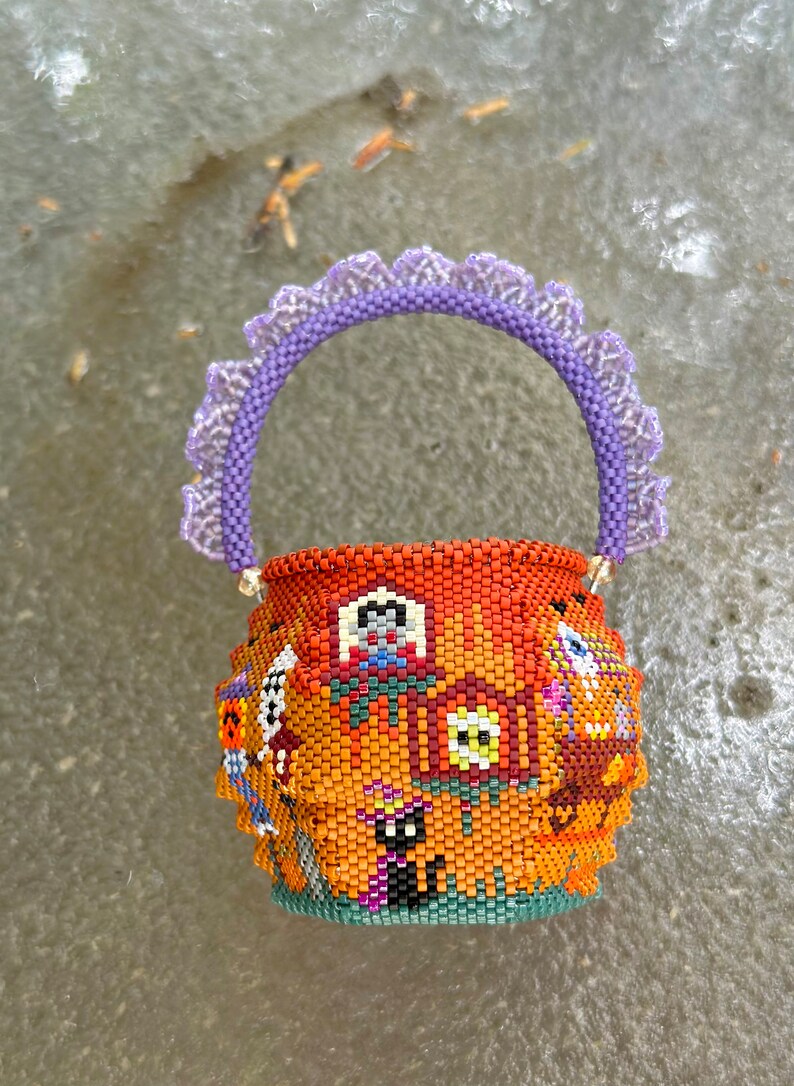 Puede incluir: Una peque&ntilde;a cesta redonda de cuentas con un fondo naranja y varios motivos coloridos de Halloween, incluyendo un gato negro, un fantasma y una casa. La cesta tiene un asa de cuentas moradas con un borde decorativo con volantes.