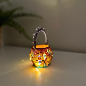 Puede incluir: Una peque&ntilde;a cesta redonda de cuentas con asa, iluminada desde el interior. El exterior presenta un colorido patr&oacute;n de Halloween con una calabaza, un fantasma y una l&aacute;pida sobre un fondo naranja. El asa tiene un borde festoneado.