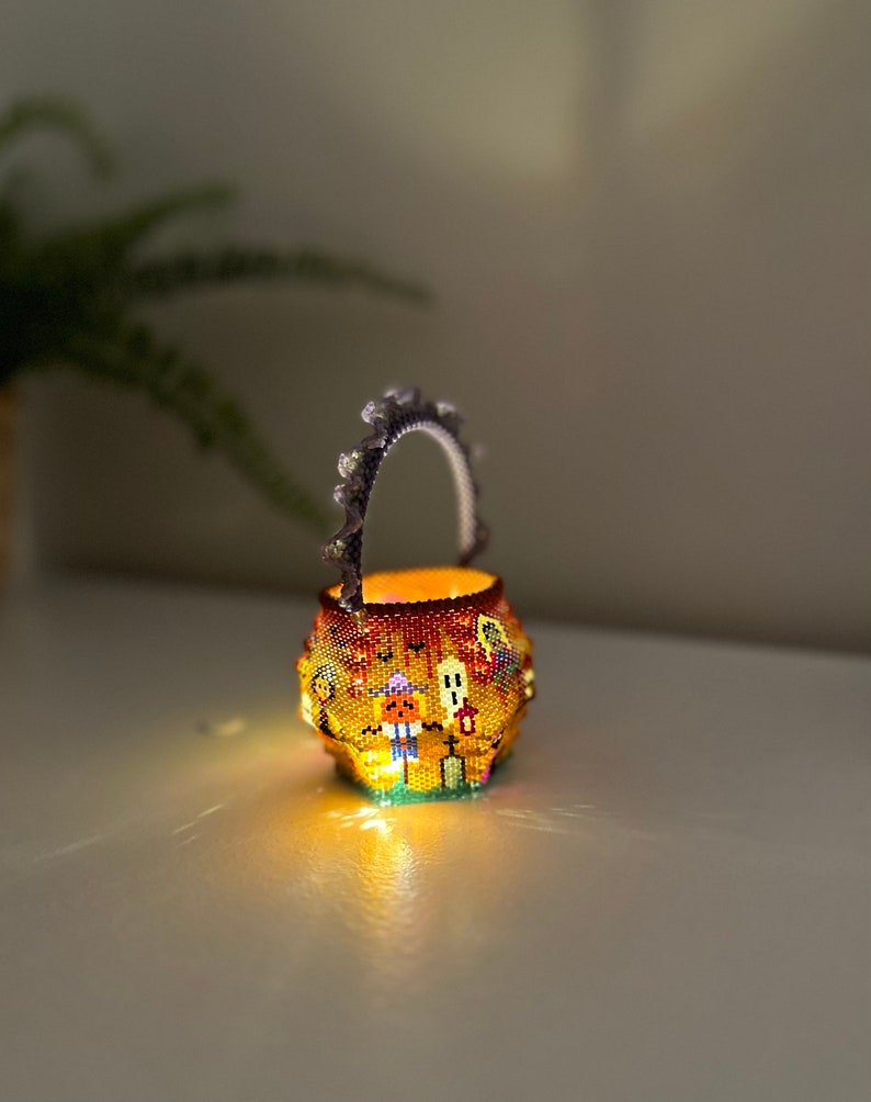 Puede incluir: Una peque&ntilde;a cesta redonda de cuentas con asa, iluminada desde el interior. El exterior presenta un colorido patr&oacute;n de Halloween con una calabaza, un fantasma y una l&aacute;pida sobre un fondo naranja. El asa tiene un borde festoneado.