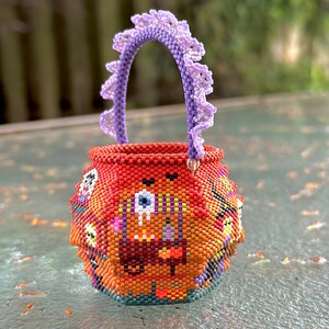Puede incluir: Una peque&ntilde;a cesta redonda de cuentas tejidas con una base de color naranja vibrante y patrones gr&aacute;ficos multicolores que incluyen un motivo de ojo. La cesta tiene un asa arqueada de color morado claro con un borde decorativo de cuentas.