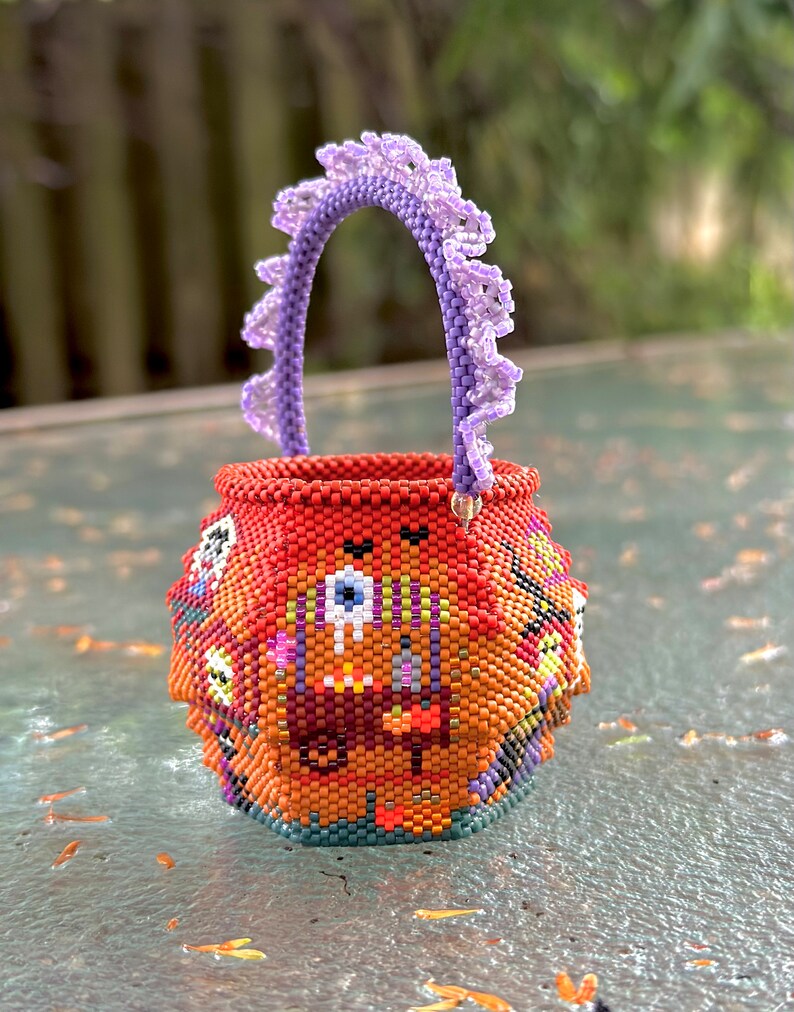 Puede incluir: Una peque&ntilde;a cesta redonda de cuentas tejidas con una base de color naranja vibrante y patrones gr&aacute;ficos multicolores que incluyen un motivo de ojo. La cesta tiene un asa arqueada de color morado claro con un borde decorativo de cuentas.