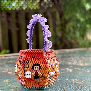 Puede incluir: Una peque&ntilde;a cesta redonda de cuentas naranjas con un patr&oacute;n detallado de un personaje, un fantasma y un bast&oacute;n. La cesta tiene un asa de cuentas moradas texturizadas y con volantes unida al borde superior, creando un recipiente decorativo.