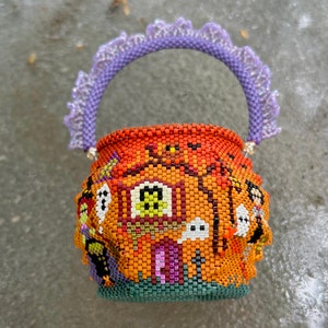 Puede incluir: Una peque&ntilde;a cesta redonda de cuentas con un dise&ntilde;o tem&aacute;tico de Halloween, que presenta un fondo naranja, una casa central, fantasmas y una bruja. La cesta tiene un asa de cuentas texturizada de color lavanda con un borde decorativo de volantes.