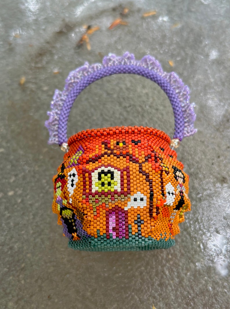 Puede incluir: Una peque&ntilde;a cesta redonda de cuentas con un dise&ntilde;o tem&aacute;tico de Halloween, que presenta un fondo naranja, una casa central, fantasmas y una bruja. La cesta tiene un asa de cuentas texturizada de color lavanda con un borde decorativo de volantes.