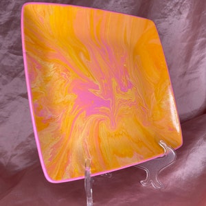 May include: A square ceramic plate with a pink and yellow marbled design. The plate is on a clear plastic stand.