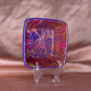 May include: A square, purple-rimmed dish with a colorful, abstract swirl design. The dish is on a clear plastic stand.