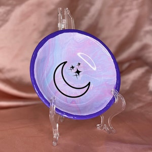May include: A small, purple-rimmed ceramic dish with a swirling pink and blue marbled design. A black crescent moon with three stars is painted in the center of the dish.