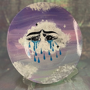 May include: A purple and white ceramic plate with a painted design of a crying cloud with blue tears. The cloud has eyelashes and a pair of eyes. The plate is on a clear plastic stand.