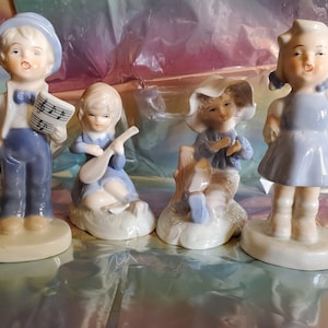 May include: Four porcelain figurines of children dressed in blue and white clothing. The figurines are standing on individual bases and are holding musical instruments.