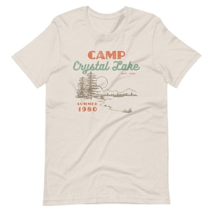 May include: A beige t-shirt with a vintage-style graphic design that reads "Camp Crystal Lake Est. 1935 Summer 1980". The graphic features a lake, mountains, and trees in a retro color palette.