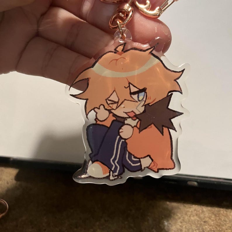 May include: A clear acrylic keychain featuring a cartoon character with orange hair, a blue and white striped shirt, and a black jacket. The character is winking and has a playful expression.