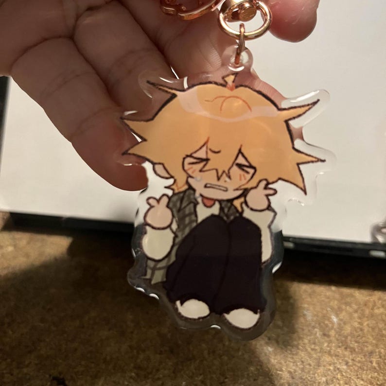 May include: A clear acrylic keychain featuring a cartoon character with blonde hair, wearing a white shirt and black pants. The character has a distressed expression and is holding their hands up to their face.