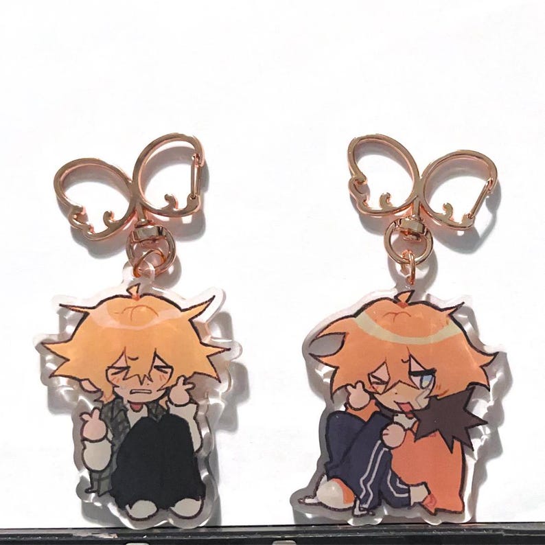 May include: Two clear acrylic keychains with gold metal rings. Each keychain features a cartoon illustration of a young person with orange hair. One keychain shows the person with their eyes closed and a worried expression, while the other keychain shows the person with one eye closed and a mischievous expression.