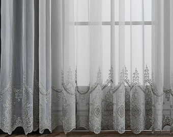 May include: White sheer curtains with an embroidered lace trim. The curtains are hanging in front of a window with a view of a city skyline. The curtains are hung on a black wall with a black picture frame hanging above them.
