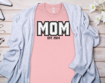 May include: Pink t-shirt with a black and white graphic that says "MOM EST. 2024".