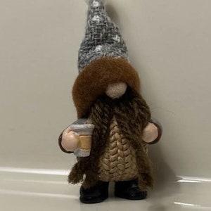 May include: A small gnome figurine wearing a gray and white knit hat with a white pom pom, a brown knit sweater, and brown boots. The gnome is holding a small cup in its hand.
