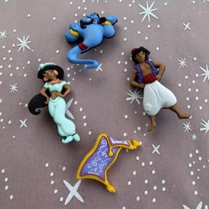 May include: Four colorful character figurines from the animated film Aladdin. The figures include the Genie, Princess Jasmine, Aladdin, and a magic carpet. The Genie is blue, Aladdin wears a red vest, and Princess Jasmine is in a teal outfit.
