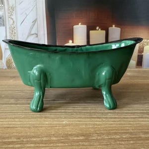 May include: A small, green bathtub-shaped decorative container with black trim. The container has four claw feet and is made of a glossy material. The background includes candles and a wooden surface.