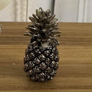 May include: A silver-colored pineapple-shaped decorative object. The pineapple has a textured surface and is sitting on a wooden surface. The pineapple's crown is detailed with individual leaves.