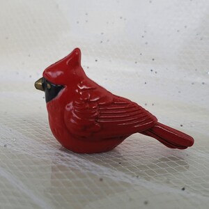 May include: A small, red cardinal figurine with black eyes and a gold beak. The figurine is made of ceramic and has a glossy finish.