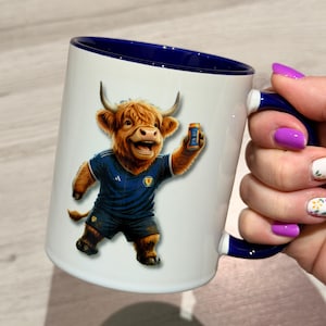 May include: White ceramic mug with a blue interior and handle. The mug features a cartoon Highland cow in a blue football kit, holding an Irn-Bru can. The mug is held by a hand with purple nail polish.