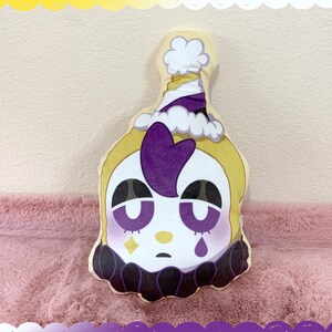 May include: A plush pillow shaped like a clown's head with a purple and white hat, yellow skin, purple hair, and a white face with black eyes, a diamond, and a tear.
