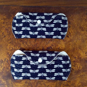 Puede incluir: Dos estuches para gafas azul marino con estampados de libélulas blancas. Cada estuche tiene un interior blanco y un cierre de botón a presión. Los estuches se muestran sobre una superficie de madera.