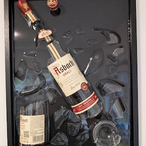 Peut inclure: Une œuvre encadrée représentant une bouteille de brandy Asbach Uralt brisée. La bouteille, avec son étiquette et ses détails rouges, est en morceaux sur un fond noir brillant. Le cadre est noir.