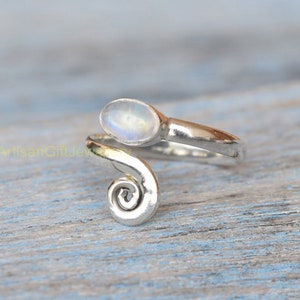 Może przedstawiać: Srebrny pierścionek z zakręconą obręczą i owalnym białym kamieniem księżycowym.