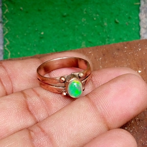 May include: A copper-colored ring with an oval, iridescent opal gemstone. The ring has a double band design with small, decorative spheres. The opal displays shades of green and yellow.