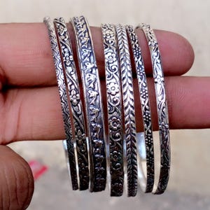 Peut inclure: Ensemble de bracelets en argent ornés de motifs floraux et de feuilles détaillés. Les bracelets sont présentés sur une main, mettant en valeur les motifs en relief. La collection présente une variété de textures, créant un ensemble de bijoux élégants.