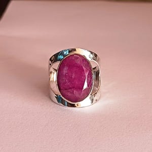 May include: A silver ring with a large, oval-shaped, pink ruby gemstone set in a wide band. The band has an open design with a cutout on either side of the stone.