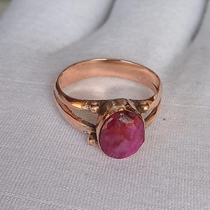 Puede incluir: Un anillo de cobre con una piedra ovalada rosa engastada en el centro. El anillo tiene un diseño de doble banda con un detalle delicado y retorcido.
