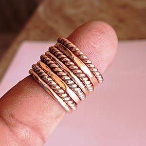 K&ouml;nnte beinhalten: Ein Satz von sieben d&uuml;nnen, goldfarbenen Ringen, die auf einem Finger gestapelt sind. Die Ringe sind alle in verschiedenen Stilen, einige sind schlicht und andere haben eine seilartige Textur.