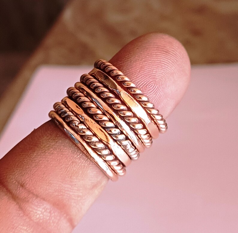 K&ouml;nnte beinhalten: Ein Satz von sieben d&uuml;nnen, goldfarbenen Ringen, die auf einem Finger gestapelt sind. Die Ringe sind alle in verschiedenen Stilen, einige sind schlicht und andere haben eine seilartige Textur.