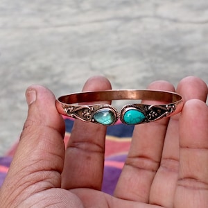 Puede incluir: Brazalete de puño color cobre con detalles ornamentados y dos piedras preciosas en forma de lágrima. Una gema es verde azulado, la otra turquesa. El brazalete se sostiene en una mano, mostrando su diseño y color.