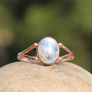 May include: A rose gold ring with a white oval moonstone gemstone. The ring has a simple design with two curved bands that meet at the top and bottom of the stone.