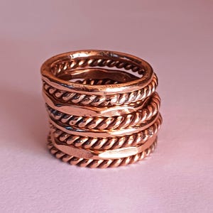 K&ouml;nnte beinhalten: Ein Stapel aus f&uuml;nf Kupferringen, jeder mit einem anderen strukturierten Design. Die Ringe sind &uuml;bereinander gestapelt und bilden ein einzigartiges und auff&auml;lliges Schmuckst&uuml;ck.