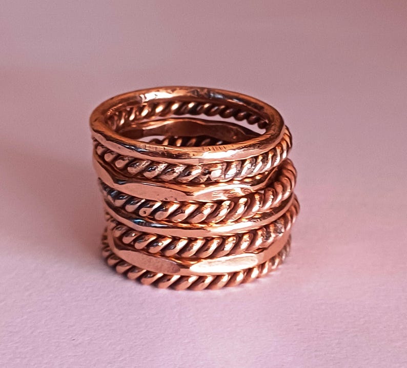 K&ouml;nnte beinhalten: Ein Stapel aus f&uuml;nf Kupferringen, jeder mit einem anderen strukturierten Design. Die Ringe sind &uuml;bereinander gestapelt und bilden ein einzigartiges und auff&auml;lliges Schmuckst&uuml;ck.