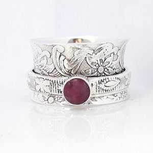 May include: A silver ring with a red gemstone and intricate floral designs. The ring is a spinner style, with a band that rotates around a central stone.
