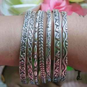 Op de afbeelding: Een set van negen zilveren armbanden met ingewikkelde bloem- en geometrische ontwerpen. De armbanden zijn gestapeld op de pols van een persoon.