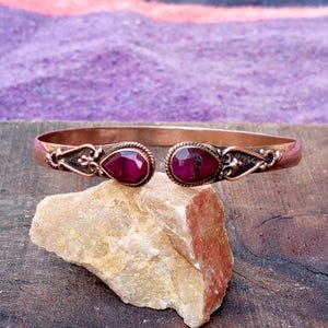 May include: A copper-colored cuff bracelet with two teardrop-shaped, deep red gemstones. The bracelet has ornate detailing near the gemstones and is displayed on a light-colored rock against a purple and gray background.